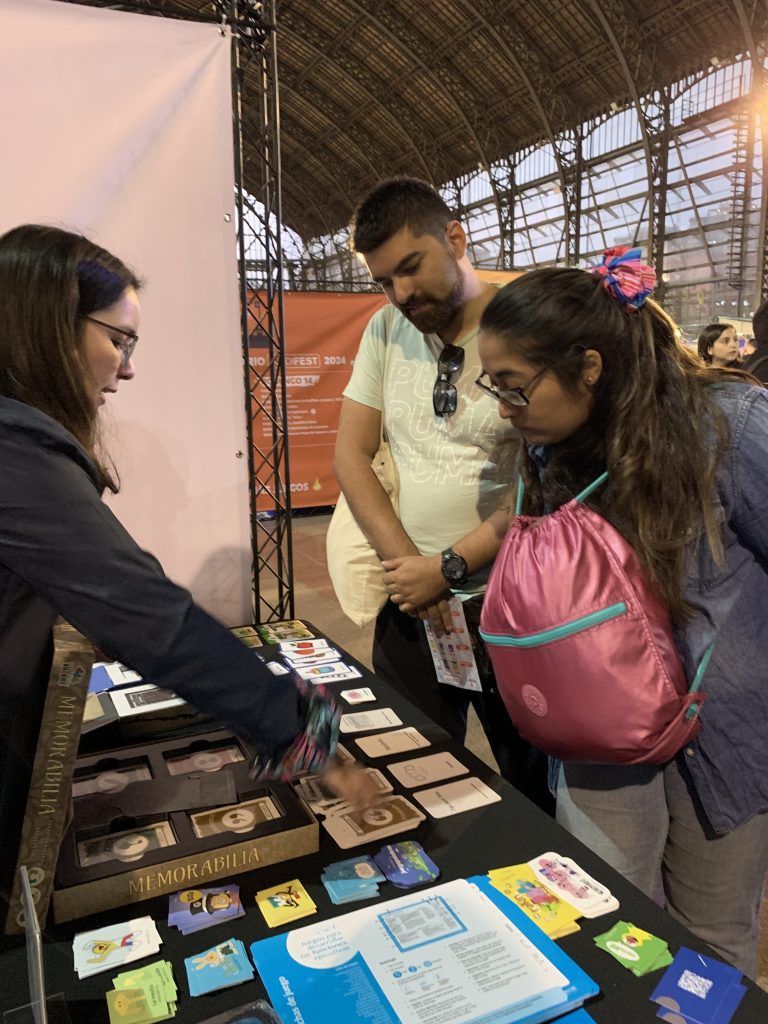 fotografía en la que aparece el stand de CEDETi UC Juegos, en el que se encuentran un hombre y una mujer, observando los juegos que se encuentran sobre la mesa del stand. Al lado contrario de la mesa, está una de la integrante de CEDETi, mostrando el juego Memorabilia. 