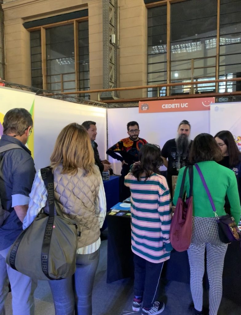 fotografía en donde aparece un grupo de cuatro personas del público, poniendo atención al stand de CEDETI UC Juegos, en el que hay cuatro integrantes del equipo de CEDETi, todos se encuentran de pie. 