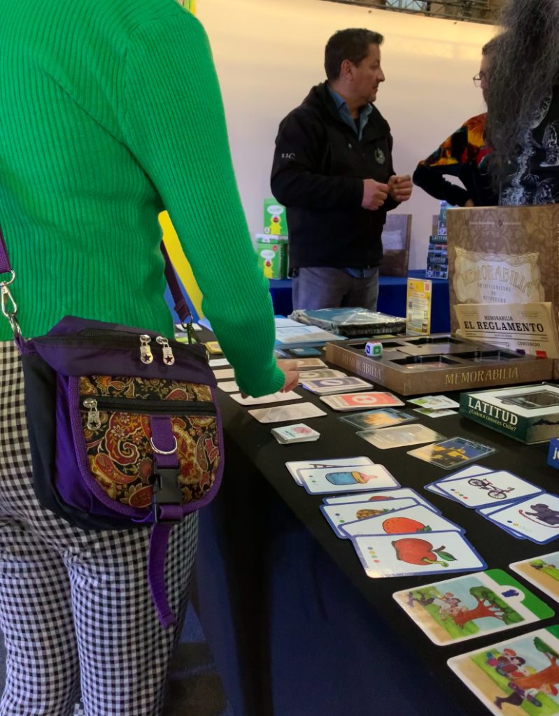 fotografía de la mesa del stand de CEDETi UC Juegos, sobre la que se encuentran las cartas de los juegos dispuestos para que el público los pueda ver. Una persona está observando las cartas, y del otro lado de la mesa, está una de las personas del equipo de CEDETi.