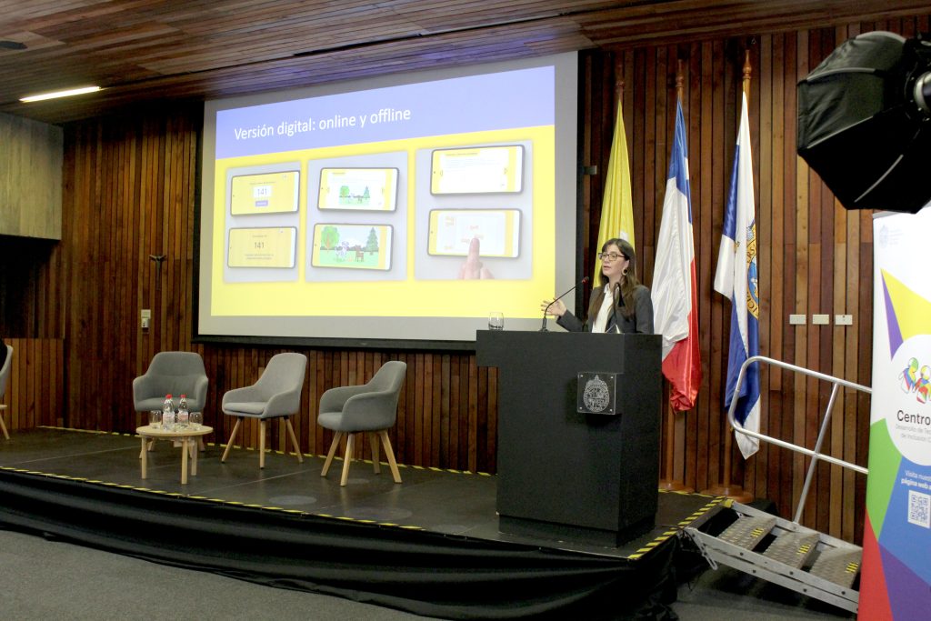 Fotografía de presentación del lanzamiento de MARKO-D. En la pared, hay una pantalla de proyección con contenido de MARKO-D. Sobre el escenario hay tres sillas ubicadas, con una mesa al centro y botellas de agua encima. Al lado de la pantalla de proyección están el podio, donde se encuentra la directora de investigación de CEDETi UC, Victoria Espinoza, exponiendo.
