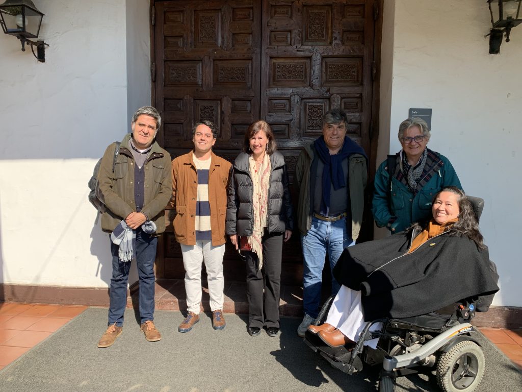 Fotografía en la que aparecen 6 personas mirando hacía la cámara. Una de ellas es usuaria de silla de ruedas y de fondo se encuentra parte de la Facultad de la escuela de Arquitectura UC