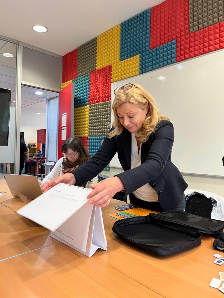 Fotografía en la que aparece la cocreadora de MARKO-D Annemarie Fritz, sosteniendo un calendario de cartón blanco, que está sobre una mesa larga de madera. A la izquierda de la foto, se encuentra sentada frente a un computador portátil la directora de Investigación de CEDETi UC, Victoria Espinoza. Atrás de ambas, se encuentra una pizarra en un muro. Sobre la mesa, también hay una funda negra, y al lado de esta, se encuentra un papel de MARKO-D