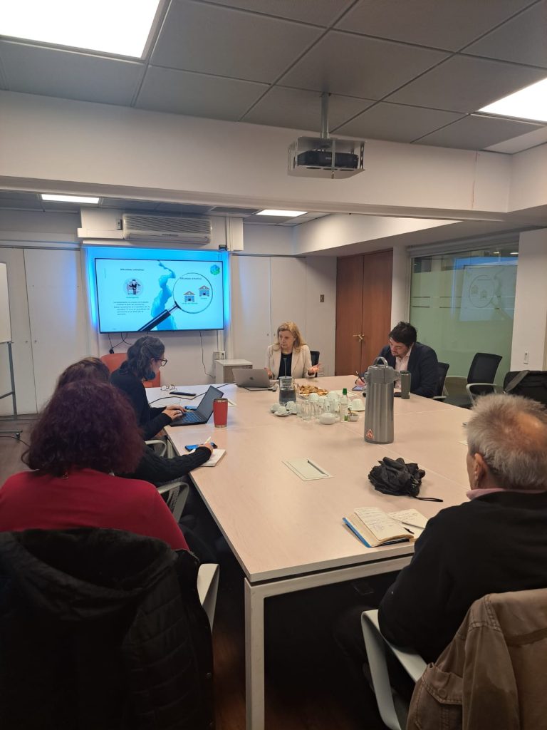Fotografía tomada desde el fondo de una sala de reuniones en la Subsecretaría de Educación Parvularia. Al centro, hay una mesa blanca de reuniones, en la que se encuentran sentados autoridades de la subsecretaría junto a la cocreadora de MARKO-D, Annemarie Fritz. La parte frontal de la sala, tiene una pizarra en la que se proyectan imágenes desde un computador.