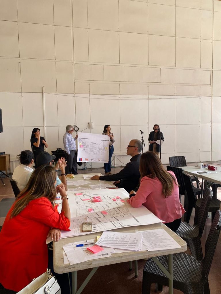 Fotografíad desde la parte de atrás de un salón en donde se ve a un grupo de personas sentadas en una mesa y otro grupo de personas exponiendo en la parte delantera.